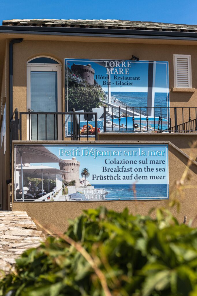 Beige building with two large signs advertising Torre Mare hotel, restaurant, and breakfast on the sea in four languages, above a stone pathway and greenery.